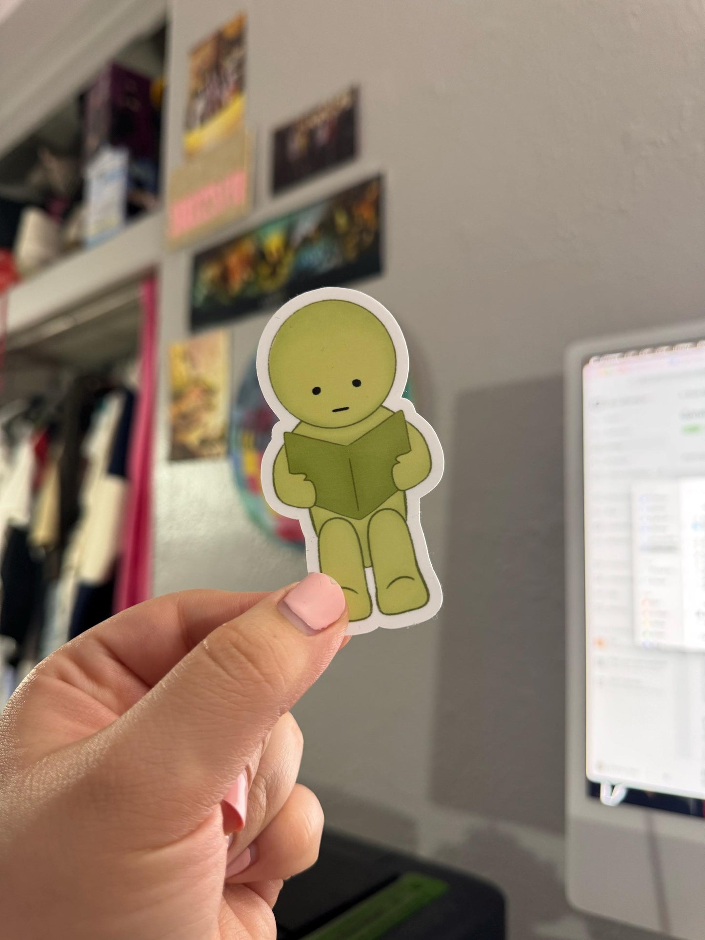 Hand holding a green cartoon character sticker in front of a blurred background